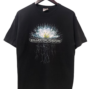 Può includere: T-shirt nera con un design floreale bianco e blu e la scritta "KILLSWITCH ENGAGE" in un ovale bianco. La maglietta è appesa a una gruccia di legno.
