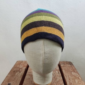Peut inclure: Un bonnet à rayures multicolores dans les tons de bleu, violet, vert, jaune et marron. Le chapeau est présenté sur une tête de mannequin contre un mur blanc et une surface en bois.