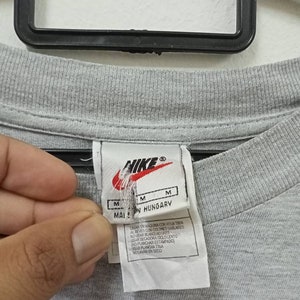 May include: Close-up of a gray t-shirt with a Nike tag. The tag includes the Nike logo, size indicators, and text that reads "MADE IN HUNGARY". Washing instructions are also printed on the tag.
