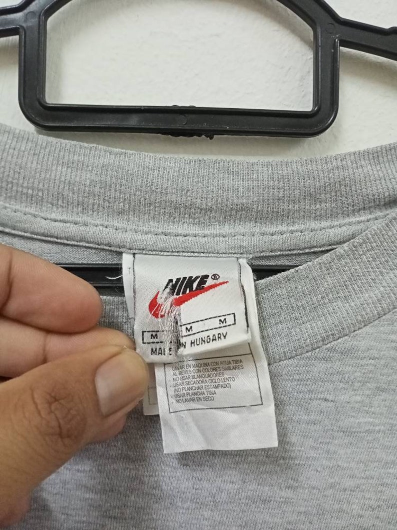May include: Close-up of a gray t-shirt with a Nike tag. The tag includes the Nike logo, size indicators, and text that reads "MADE IN HUNGARY". Washing instructions are also printed on the tag.