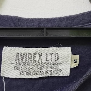 K&ouml;nnte beinhalten: Nahaufnahme eines marineblauen T-Shirt-Etiketts. Auf dem Etikett steht "AVIREX LTD" mit zus&auml;tzlichem Text: "ARMED FORCES MANUFACTURER". Das Etikett enth&auml;lt auch die Gr&ouml;&szlig;e "M" und den Text "ESTABLISHED IN 1975".