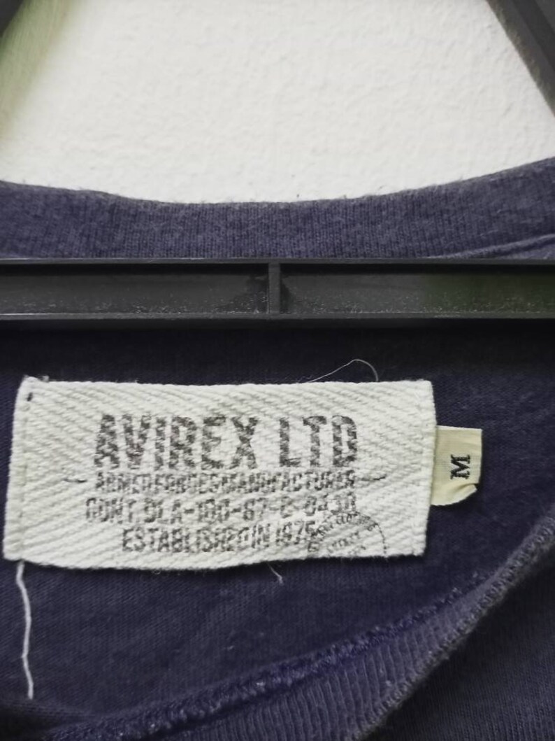 K&ouml;nnte beinhalten: Nahaufnahme eines marineblauen T-Shirt-Etiketts. Auf dem Etikett steht "AVIREX LTD" mit zus&auml;tzlichem Text: "ARMED FORCES MANUFACTURER". Das Etikett enth&auml;lt auch die Gr&ouml;&szlig;e "M" und den Text "ESTABLISHED IN 1975".