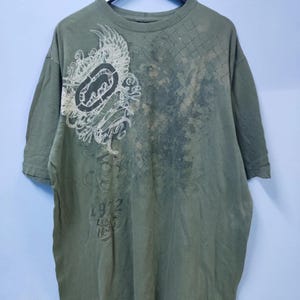 Puede incluir: Camiseta verde oliva con un gran diseño gráfico en la parte delantera. El diseño incluye un logotipo estilizado y elementos decorativos en tonos blancos y grises. La camiseta también presenta el texto "1972" y "tek" en una fuente desgastada.