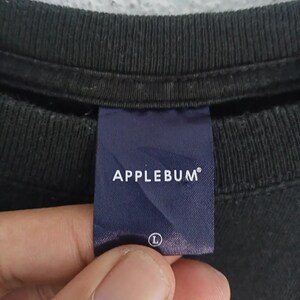 K&ouml;nnte beinhalten: Nahaufnahme eines schwarzen T-Shirt-Kragens mit einem marineblauen Etikett. Auf dem Etikett steht "APPLEBUM" in wei&szlig;en Gro&szlig;buchstaben. Auf dem Etikett ist die Gr&ouml;&szlig;e "L" angegeben. Das Shirt ist aus dunklem Stoff.