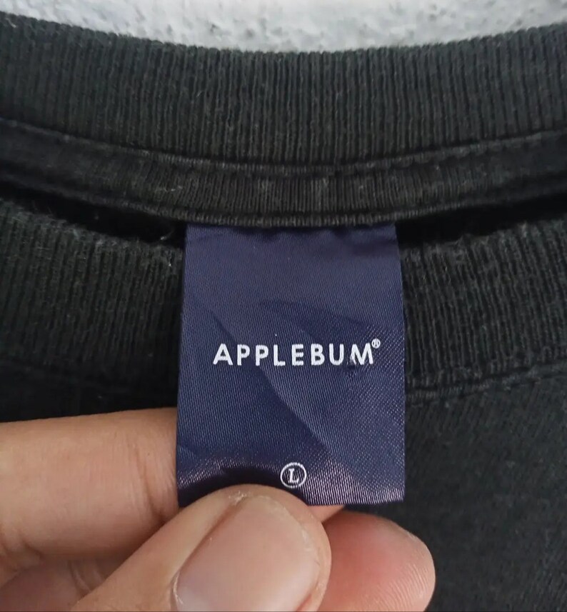 K&ouml;nnte beinhalten: Nahaufnahme eines schwarzen T-Shirt-Kragens mit einem marineblauen Etikett. Auf dem Etikett steht "APPLEBUM" in wei&szlig;en Gro&szlig;buchstaben. Auf dem Etikett ist die Gr&ouml;&szlig;e "L" angegeben. Das Shirt ist aus dunklem Stoff.