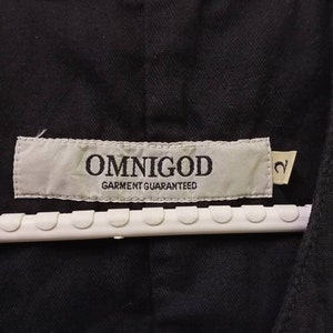 Op de afbeelding: Een zwart kledingstuk met een wit label waarop "OMNIGOD GARMENT GUARANTEED" staat en een maatlabel met de tekst "2".