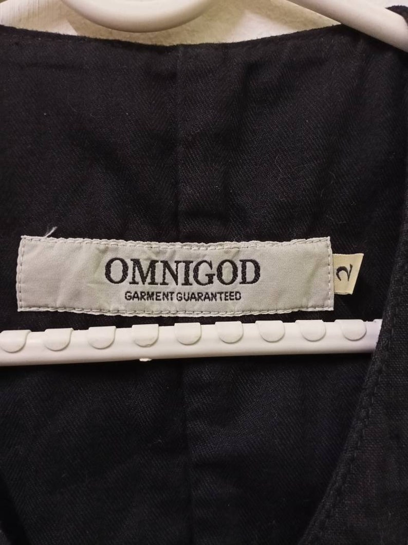 Op de afbeelding: Een zwart kledingstuk met een wit label waarop "OMNIGOD GARMENT GUARANTEED" staat en een maatlabel met de tekst "2".