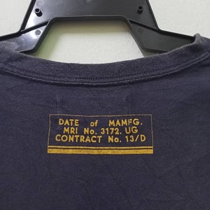 K&ouml;nnte beinhalten: Marineblaues T-Shirt mit einem goldenen rechteckigen Etikett auf der R&uuml;ckseite. Das Etikett enth&auml;lt Text wie "DATE of MAMFG", "MRI No. 3172. UG" und "CONTRACT No. 13/D". Das T-Shirt h&auml;ngt an einem schwarzen Kleiderb&uuml;gel.