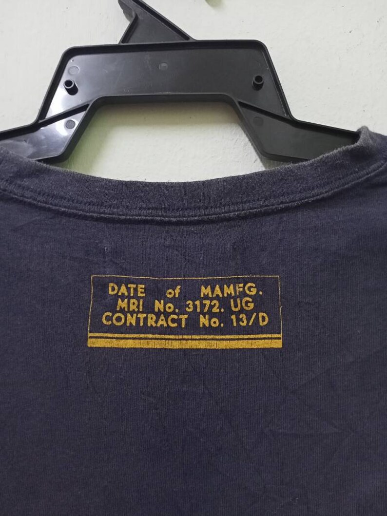 K&ouml;nnte beinhalten: Marineblaues T-Shirt mit einem goldenen rechteckigen Etikett auf der R&uuml;ckseite. Das Etikett enth&auml;lt Text wie "DATE of MAMFG", "MRI No. 3172. UG" und "CONTRACT No. 13/D". Das T-Shirt h&auml;ngt an einem schwarzen Kleiderb&uuml;gel.