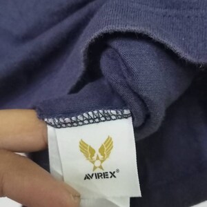 K&ouml;nnte beinhalten: Nahaufnahme eines marineblauen T-Shirts mit einem Etikett mit dem Avirex-Logo. Das Logo zeigt goldene Fl&uuml;gel und einen Stern, darunter der Markenname. Der Stoff ist einfarbig dunkelblau.