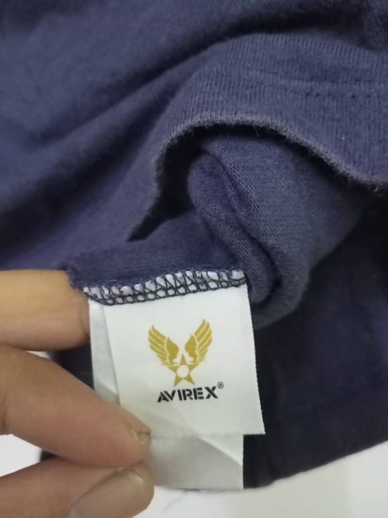 K&ouml;nnte beinhalten: Nahaufnahme eines marineblauen T-Shirts mit einem Etikett mit dem Avirex-Logo. Das Logo zeigt goldene Fl&uuml;gel und einen Stern, darunter der Markenname. Der Stoff ist einfarbig dunkelblau.