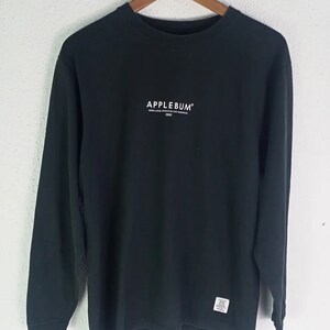 K&ouml;nnte beinhalten: Schwarzes Langarmshirt mit dem Markennamen "APPLEBUM" in Wei&szlig; auf der Brust. Das Shirt hat auch zus&auml;tzlichen Text unter dem Markennamen und ein kleines Etikett in der N&auml;he des Saums. Das Shirt h&auml;ngt an einem Holzb&uuml;gel.