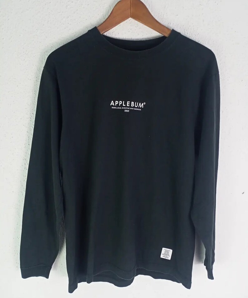 K&ouml;nnte beinhalten: Schwarzes Langarmshirt mit dem Markennamen "APPLEBUM" in Wei&szlig; auf der Brust. Das Shirt hat auch zus&auml;tzlichen Text unter dem Markennamen und ein kleines Etikett in der N&auml;he des Saums. Das Shirt h&auml;ngt an einem Holzb&uuml;gel.