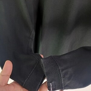 K&ouml;nnte beinhalten: Schwarzes Langarmshirt mit geripptem B&uuml;ndchen. Das B&uuml;ndchen hat ein kleines rechteckiges Etikett mit dem wei&szlig;en Text "APPLEBUM". Das Shirt ist aus einem dunklen Stoff gefertigt.