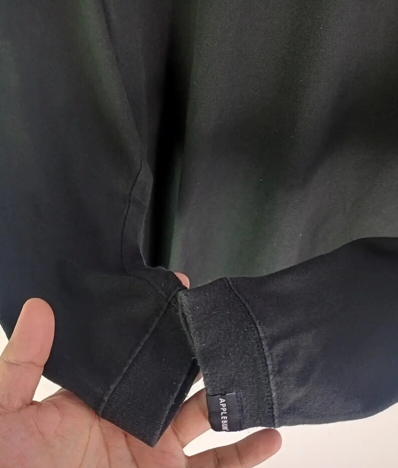 K&ouml;nnte beinhalten: Schwarzes Langarmshirt mit geripptem B&uuml;ndchen. Das B&uuml;ndchen hat ein kleines rechteckiges Etikett mit dem wei&szlig;en Text "APPLEBUM". Das Shirt ist aus einem dunklen Stoff gefertigt.