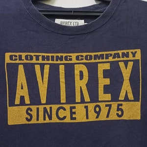 K&ouml;nnte beinhalten: Marineblaues T-Shirt mit einem goldenen rechteckigen Aufdruck mit der Aufschrift "CLOTHING COMPANY AVIREX SINCE 1975". Das T-Shirt hat einen Rundhalsausschnitt und h&auml;ngt an einem schwarzen Kleiderb&uuml;gel. Der Markenname "AVIREX LTD" ist auf einem Etikett aufgedruckt.