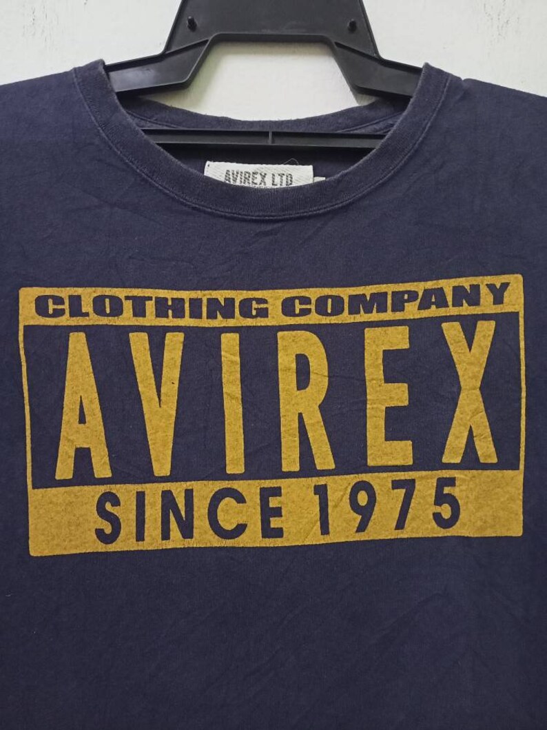 K&ouml;nnte beinhalten: Marineblaues T-Shirt mit einem goldenen rechteckigen Aufdruck mit der Aufschrift "CLOTHING COMPANY AVIREX SINCE 1975". Das T-Shirt hat einen Rundhalsausschnitt und h&auml;ngt an einem schwarzen Kleiderb&uuml;gel. Der Markenname "AVIREX LTD" ist auf einem Etikett aufgedruckt.