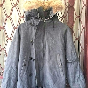 Könnte beinhalten: Grauer Winterparka mit fellgefütterter Kapuze. Die Jacke hat einen Reißverschluss, eine Knopfleiste und mehrere Taschen. Der Fellbesatz ist hellbraun.