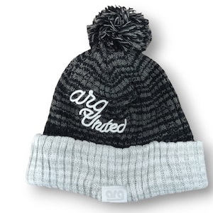 May include: A black and gray knit beanie with a gray ribbed cuff and a matching pom-pom. The white script text "arg United" is embroidered on the front. A small white tag with the "arg" logo is sewn on the cuff.