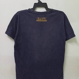 K&ouml;nnte beinhalten: Marineblaues T-Shirt, das an einem schwarzen Kleiderb&uuml;gel h&auml;ngt. Auf der R&uuml;ckseite des Shirts befindet sich ein rechteckiger gelber Aufn&auml;her mit schwarzem Text. Der Text enth&auml;lt die W&ouml;rter "RATE OF MANFG" und "CONTRACT No. 13/9".