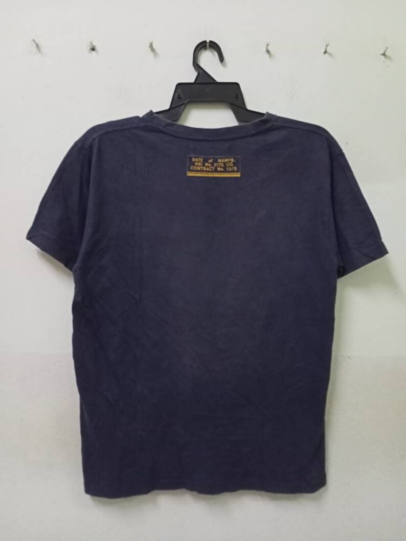 K&ouml;nnte beinhalten: Marineblaues T-Shirt, das an einem schwarzen Kleiderb&uuml;gel h&auml;ngt. Auf der R&uuml;ckseite des Shirts befindet sich ein rechteckiger gelber Aufn&auml;her mit schwarzem Text. Der Text enth&auml;lt die W&ouml;rter "RATE OF MANFG" und "CONTRACT No. 13/9".