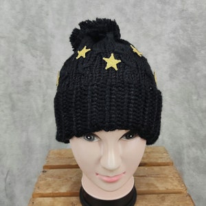 May include: A black knit beanie with a pom-pom and gold star appliqués. The hat is displayed on a mannequin head. The beanie is made of a thick, textured yarn, and the stars are evenly spaced across the top.