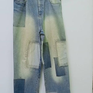 Könnte beinhalten: Hellblaue Jeans aus Denim mit einem Patchwork-Design in verschiedenen Blau- und Weißtönen. Die Hose verfügt über sichtbare Ziernähte an den Flicken, einen Knopfverschluss, Gürtelschlaufen und ein schwarzes Markenetikett an der Vordertasche.