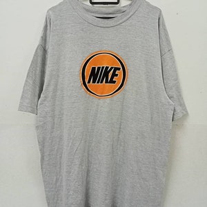 May include: A gray t-shirt with a large orange circle design in the center. The word "NIKE" is printed in black within the orange circle. The shirt is hanging on a black hanger.