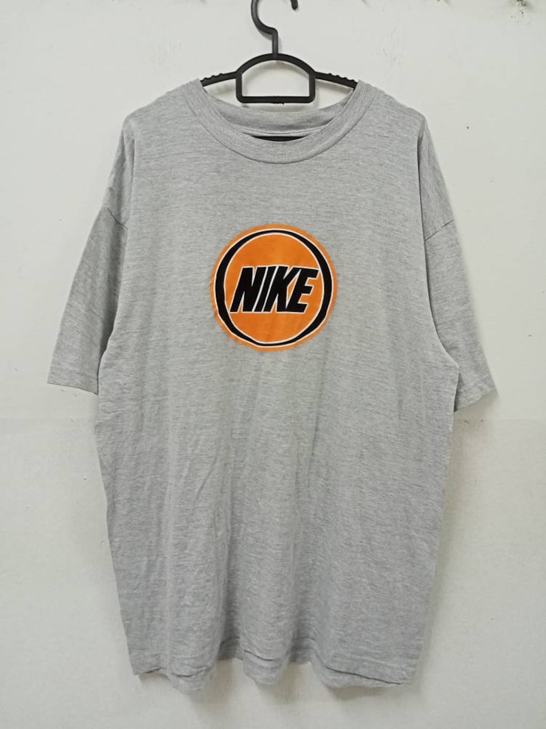 May include: A gray t-shirt with a large orange circle design in the center. The word "NIKE" is printed in black within the orange circle. The shirt is hanging on a black hanger.