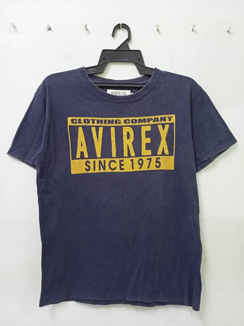 K&ouml;nnte beinhalten: Marineblaues T-Shirt mit gelbem rechteckigem Aufdruck "AVIREX CLOTHING COMPANY SINCE 1975". Das T-Shirt h&auml;ngt an einem schwarzen Kleiderb&uuml;gel. Der Markenname ist auch auf dem Kragen aufgedruckt.