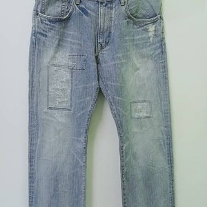 May include: Light blue denim jeans with distressed details, patches, and a classic five-pocket design. The jeans are hanging on a black hanger against a white background. The jeans have a faded wash and a relaxed fit.