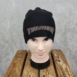 Może przedstawiać: Czarna dzianinowa czapka z napisem "THRASHER" w stylu dżetów z przodu. Czapka jest prezentowana na głowie manekina. Klasyczny dodatek na zimę.