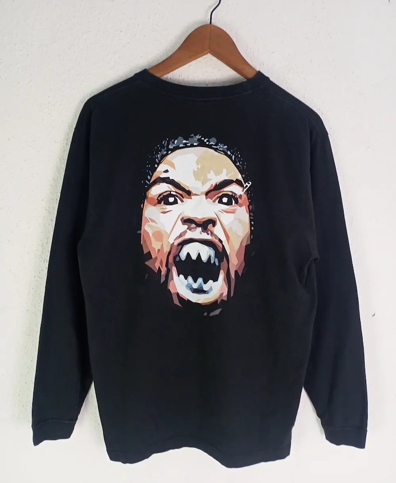 K&ouml;nnte beinhalten: Schwarzes Langarmshirt mit einem grafischen Portr&auml;t auf dem R&uuml;cken. Das Portr&auml;t ist eine stilisierte, farbenfrohe Darstellung eines Gesichts mit offenem Mund und sichtbaren Z&auml;hnen. Das Shirt h&auml;ngt an einem Holzkleiderb&uuml;gel.