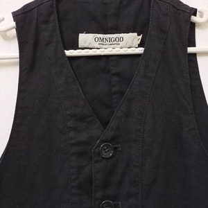 Op de afbeelding: Een zwart, mouwloos vest met een V-hals en twee knopen. Het vest heeft een zak aan de voorkant en een label met de tekst "OMNIGOD GARMENT GUARANTEED".
