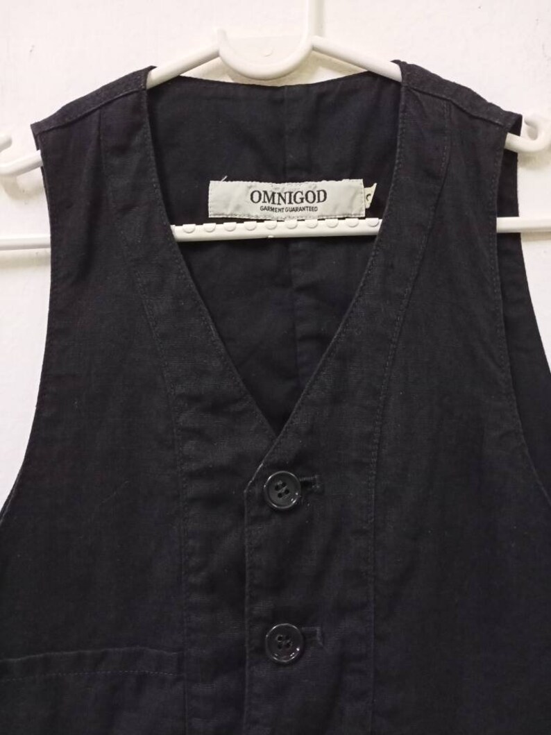 Op de afbeelding: Een zwart, mouwloos vest met een V-hals en twee knopen. Het vest heeft een zak aan de voorkant en een label met de tekst "OMNIGOD GARMENT GUARANTEED".