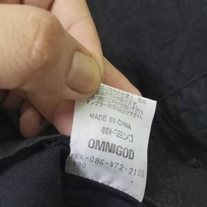 Op de afbeelding: Een wit label met zwarte tekst dat "MADE IN CHINA", "OMNIGOD" en een telefoonnummer vermeldt. Het label is bevestigd aan een zwart kledingstuk.
