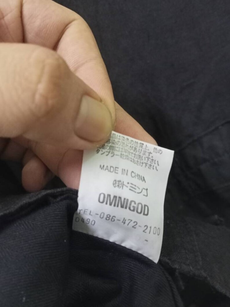 Op de afbeelding: Een wit label met zwarte tekst dat "MADE IN CHINA", "OMNIGOD" en een telefoonnummer vermeldt. Het label is bevestigd aan een zwart kledingstuk.