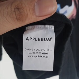 K&ouml;nnte beinhalten: Schwarzes Kleidungsst&uuml;cketikett mit dem Markennamen "APPLEBUM" in fetten wei&szlig;en Buchstaben. Darunter befinden sich japanische Schriftzeichen und Kontaktinformationen, einschlie&szlig;lich einer Telefonnummer und einer Website-Adresse.