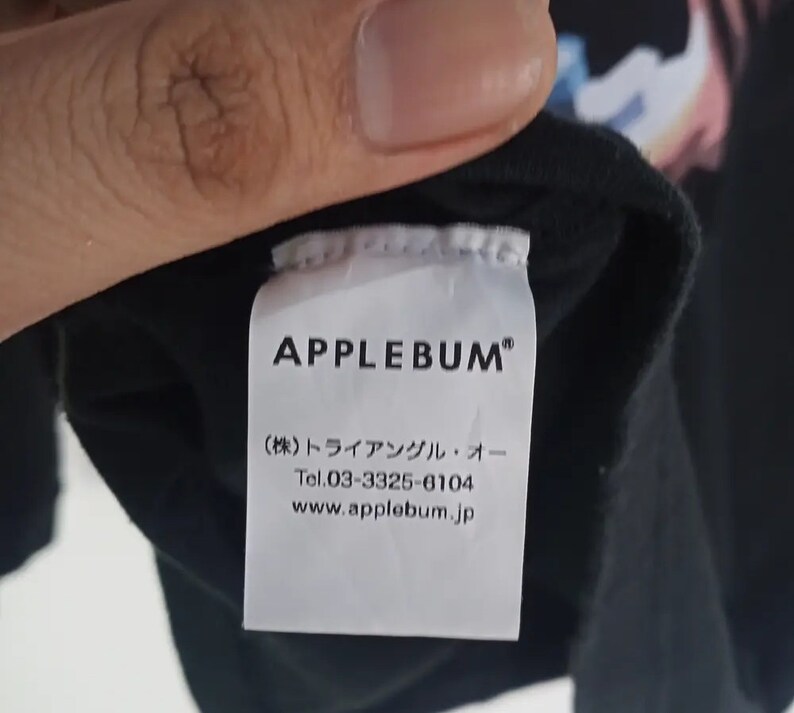 K&ouml;nnte beinhalten: Schwarzes Kleidungsst&uuml;cketikett mit dem Markennamen "APPLEBUM" in fetten wei&szlig;en Buchstaben. Darunter befinden sich japanische Schriftzeichen und Kontaktinformationen, einschlie&szlig;lich einer Telefonnummer und einer Website-Adresse.