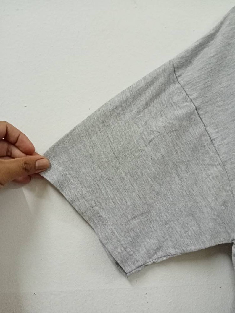 May include: Close-up of a gray t-shirt sleeve. The fabric appears to be a soft, knit material. The sleeve is a light gray color and has a simple, clean design. The sleeve is being held by a hand.