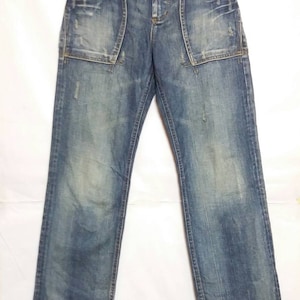 Könnte beinhalten: Blaue Jeans aus Denim mit Used-Waschung, Knopfleiste und zwei großen, geschwungenen Vordertaschen. Die Hose hat einen geradlinigen Schnitt mit sichtbaren Verblassungen an Oberschenkeln und Knien sowie verstärkten Nähten an den Taschen.