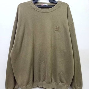 Könnte beinhalten: Olivgrünes Sweatshirt mit Rundhalsausschnitt und langen Ärmeln. Es hat ein kleines gesticktes Logo auf der Brust. Das Sweatshirt hängt an einem schwarzen Kleiderbügel vor weißem Hintergrund. Der Stoff scheint weich und bequem zu sein.