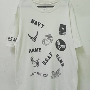 Op de afbeelding: Wit T-shirt met ronde hals en korte mouwen, versierd met militaire emblemen en tekst zoals "NAVY", "ARMY" en "U.S.A.F.". Het ontwerp bevat ankers, sterren en andere militaire insignes, gedrukt in zwart.