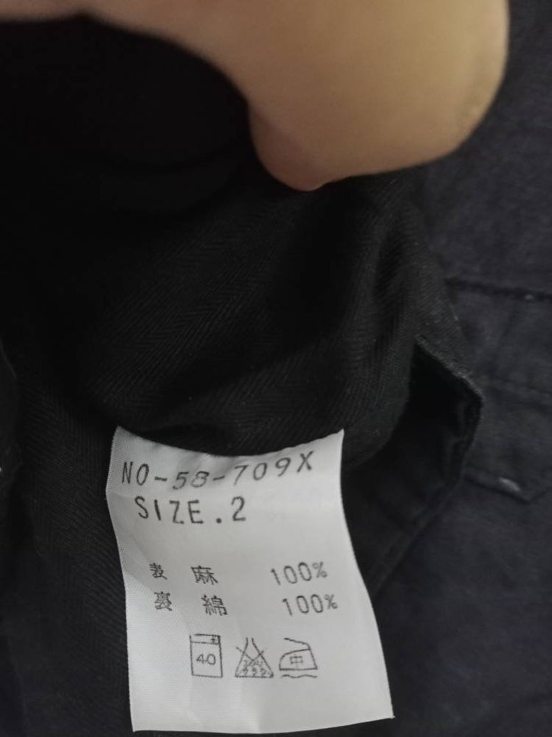 Op de afbeelding: Een zwart kledinglabel met witte tekst die "NO-58-709X SIZE. 2" en "綿 100%  綿 100%" luidt. Het label bevat ook een onderhoudsetiket met een was symbool voor 40 graden Celsius, een symbool voor verboden bleken, een strijksymbool voor lage hitte en een symbool voor verboden chemische reiniging.