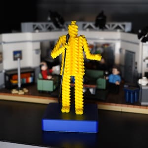 May include: A yellow Lego figure of a comedian with a microphone stands on a blue base, in front of a Lego set of a sitcom apartment. The figure is made of yellow Lego bricks. The apartment set includes furniture and other Lego figures.