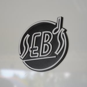 May include: A black and white emblem with the letters "SEB's" inside a circle. The emblem includes a musical note and decorative lines. The design is set against a white background.