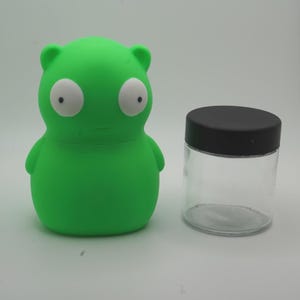 May include: A green, cartoon-like figure with large white eyes and a small mouth, next to a clear glass jar with a black lid. The figure has small arms and ears. The jar is cylindrical.