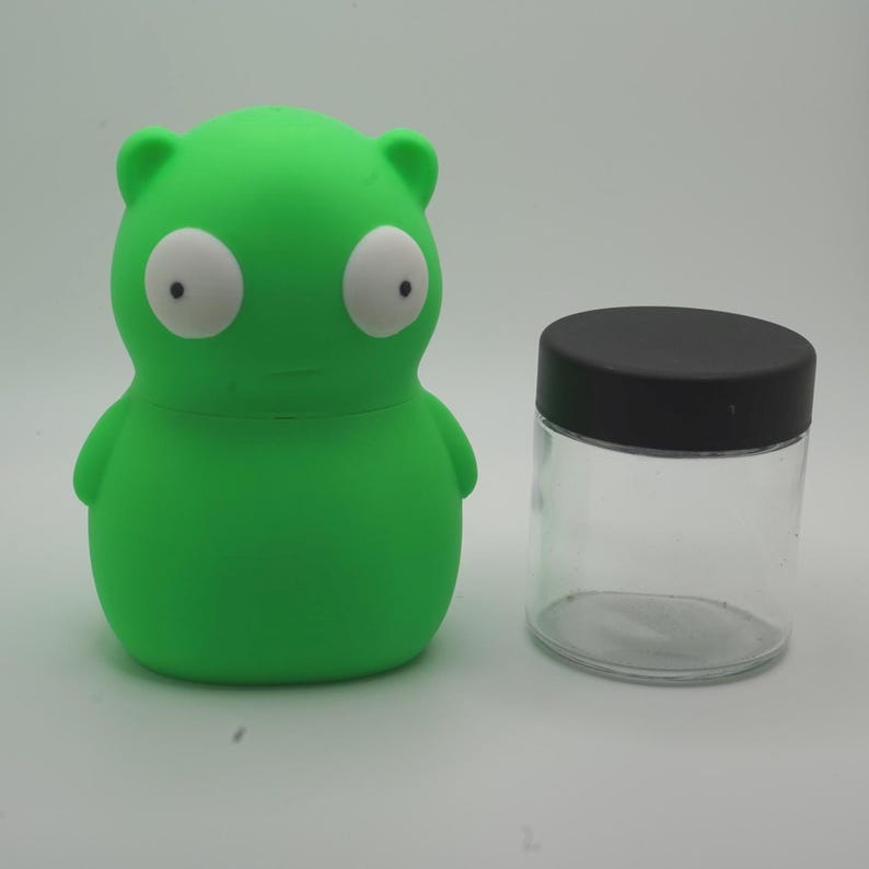 May include: A green, cartoon-like figure with large white eyes and a small mouth, next to a clear glass jar with a black lid. The figure has small arms and ears. The jar is cylindrical.