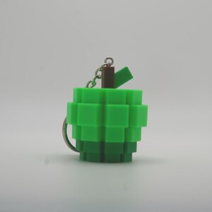 以下が含まれることがあります： 緑色のピクセル化された手榴弾型のキーホルダー。キーホルダーは、銀色のリングとチェーン、上部に茶色と緑色のアクセントが付いています。デザインはビデオゲームの美学を彷彿とさせます。