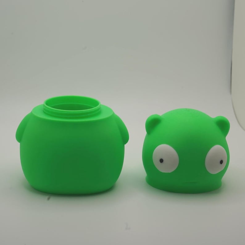 May include: Two-piece green container set. The larger piece is a round, squat shape with a wide opening and two small side handles. The smaller piece is a round lid with a cartoonish face, white eyes, and small ears.